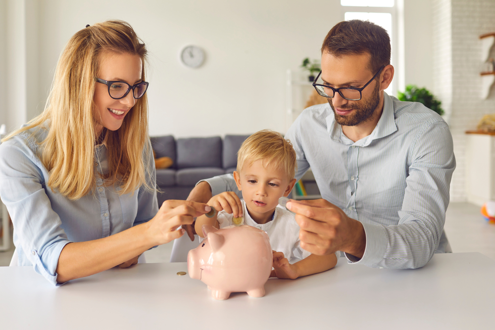 Little child puts money inside piggy bank, learns to effectively manage budget and spend wisely from young independent parents able to provide high living standard