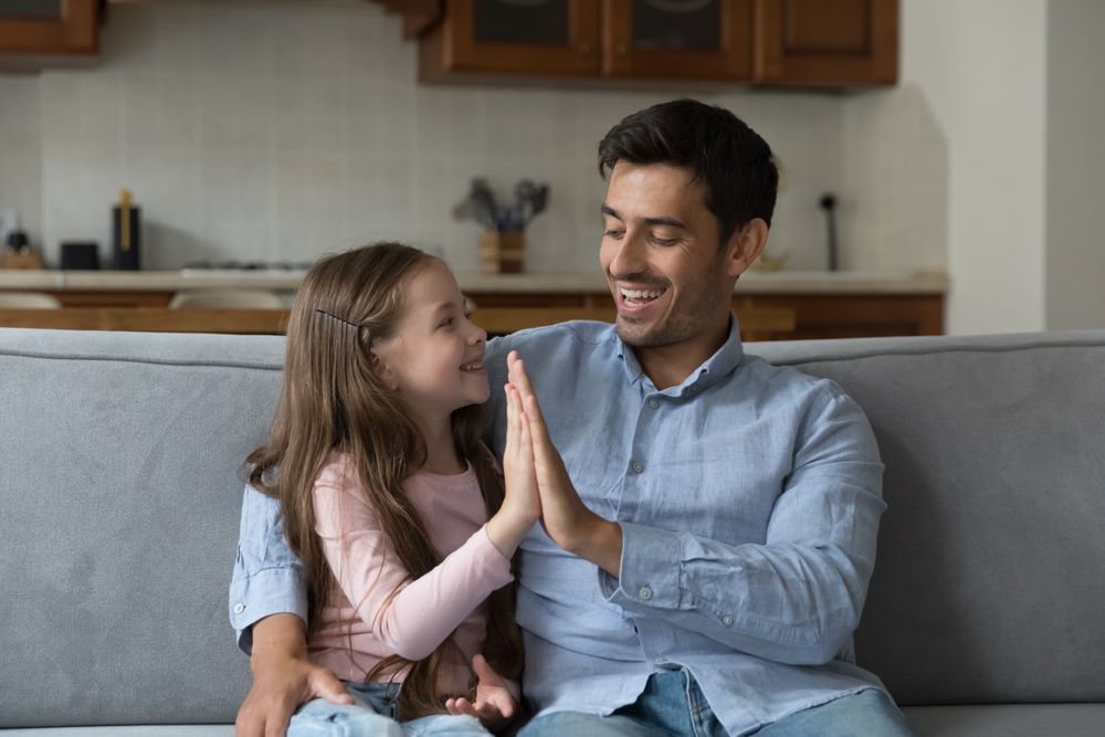 Proud kid girl and happy father giving high five on home couch, hugging, talking, laughing, enjoying leisure
