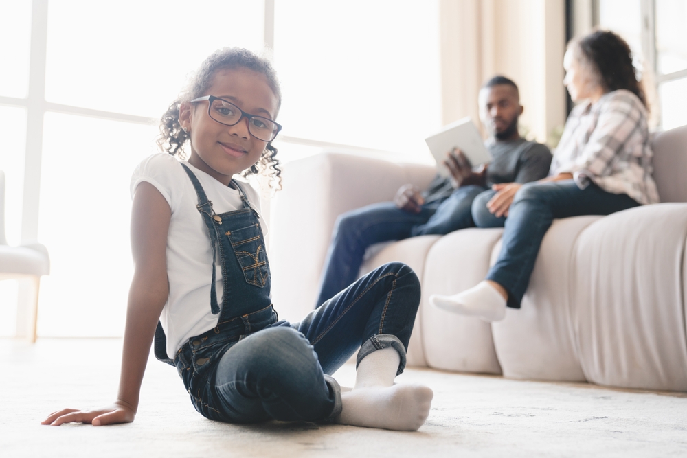 African-american child girl kid daughter sitting relaxing on the floor while parents spouses using digital tablet and watching movie, surfing social media application at home