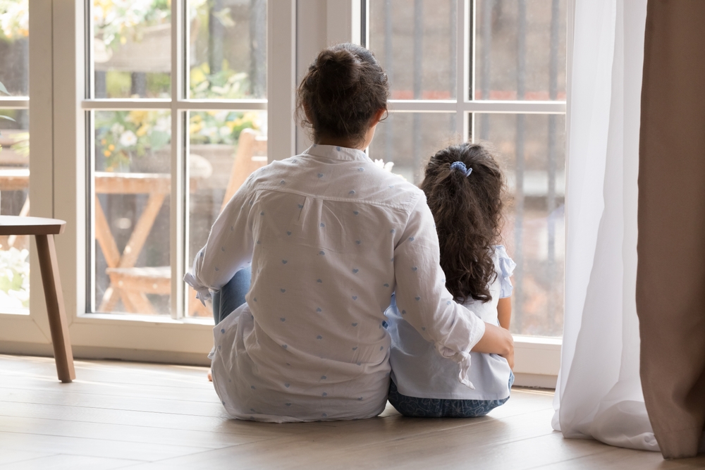 Rear view young loving mother hugging little daughter sitting on warm floor near window looking together outside